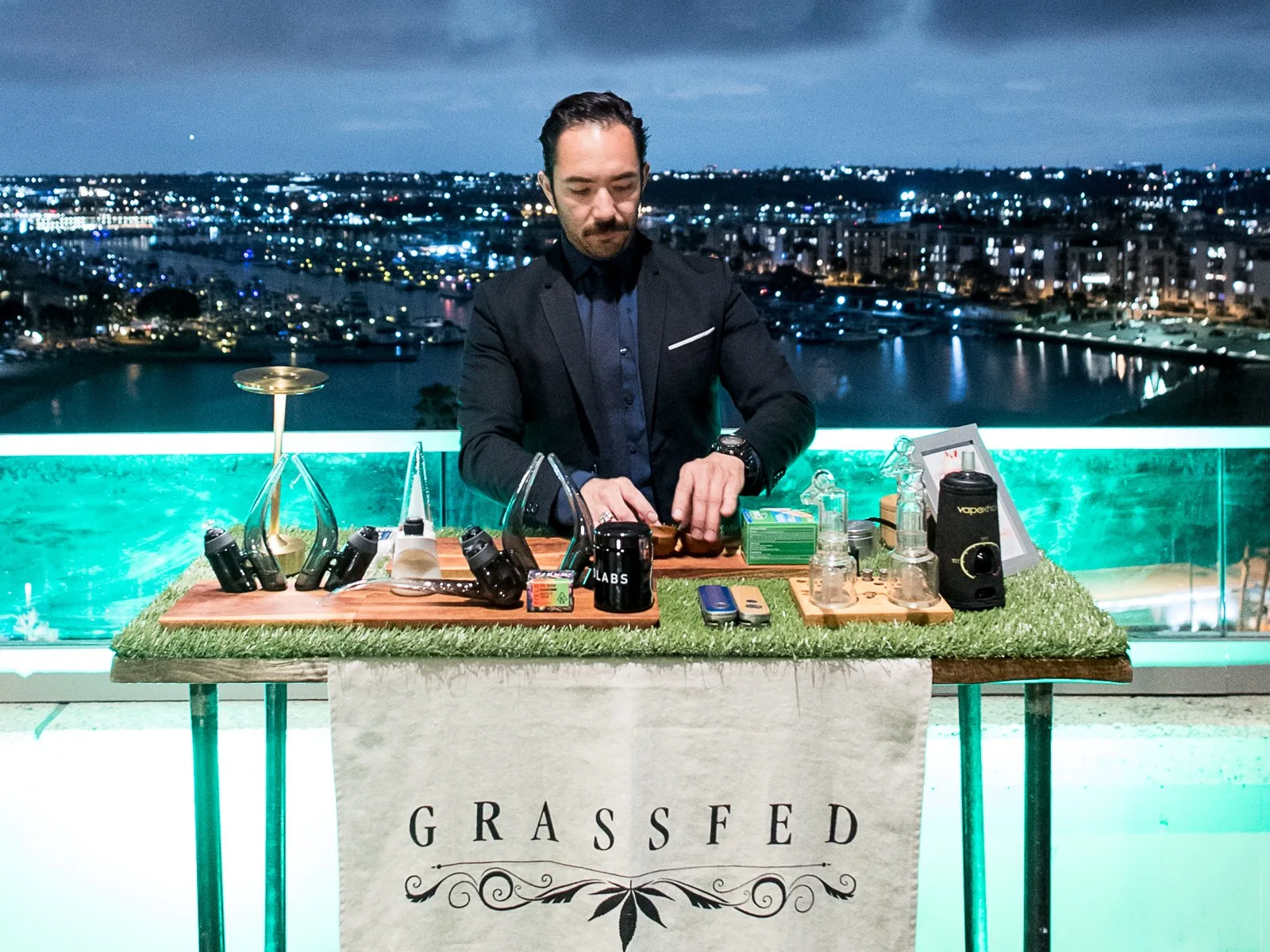 A budtender in a dark suit preparing devices behind the Grassfed cannabis bar on a rooftop at night, city lights behind him