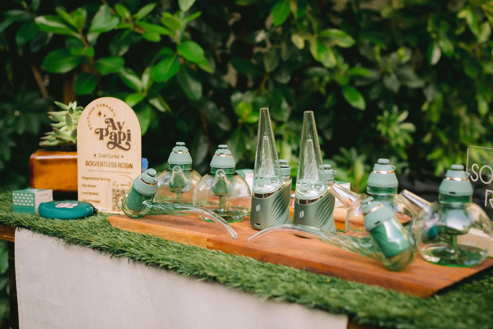 Devices and glassware lined up on a grass-topped cannabis bar in a garden, next to an Ay Papi sign