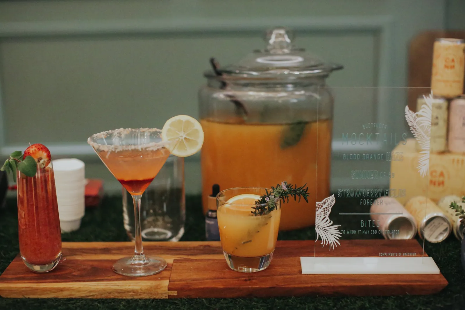 Craft mocktails garnished with strawberry, lemon and rosemary on a wood and grass bar beside a glass drink dispenser