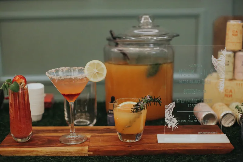 Craft mocktails garnished with strawberry, lemon and rosemary on a wood and grass bar beside a glass drink dispenser