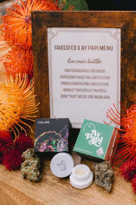 A framed Grassfed x Ay Papi menu of live rosin strains surrounded by orange protea flowers and Ay Papi jars