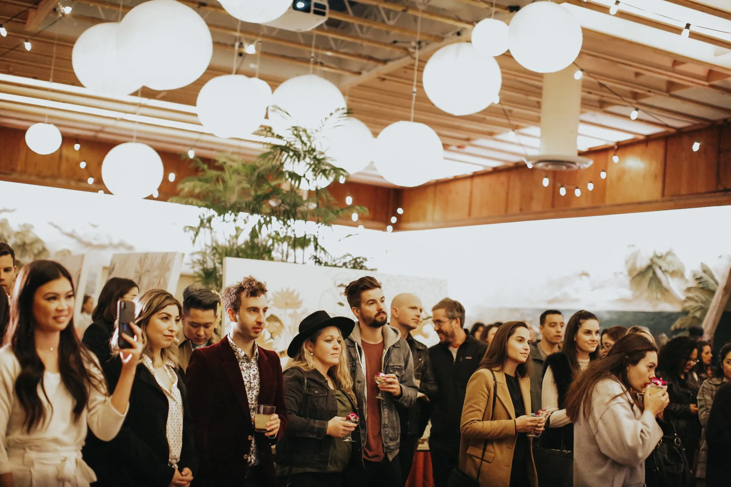 A crowd of guests with drinks under white paper lanterns at an indoor event