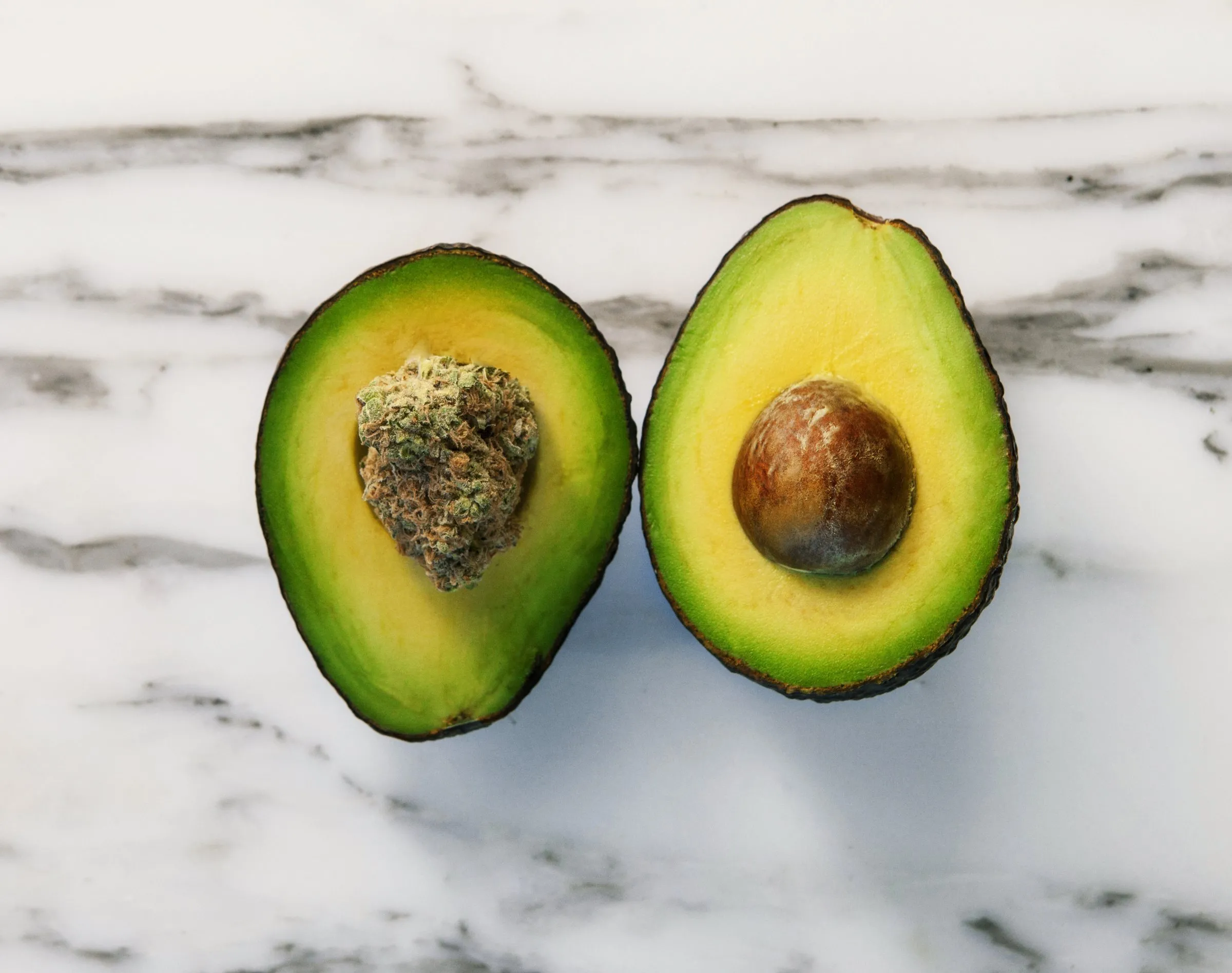 A halved avocado with a cannabis flower in place of the pit on white marble