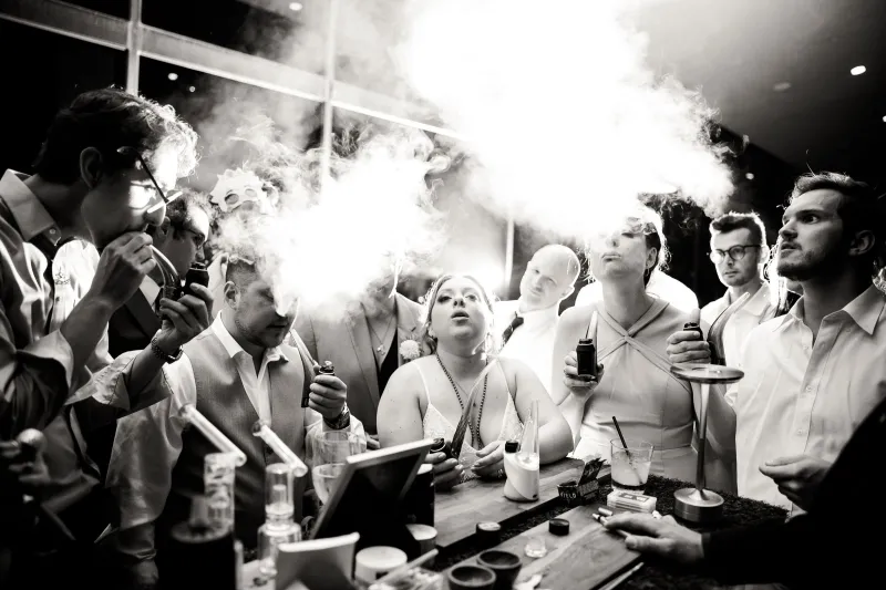 Black and white photo of wedding guests gathered around the cannabis bar, the bride in the center exhaling a cloud of vapor
