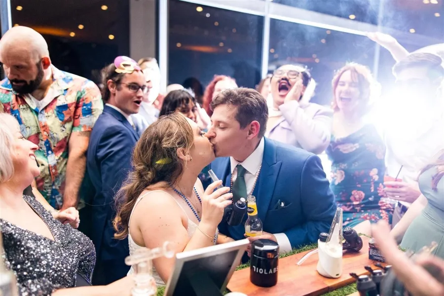 Bride and groom kissing at the cannabis bar while their wedding guests cheer behind them