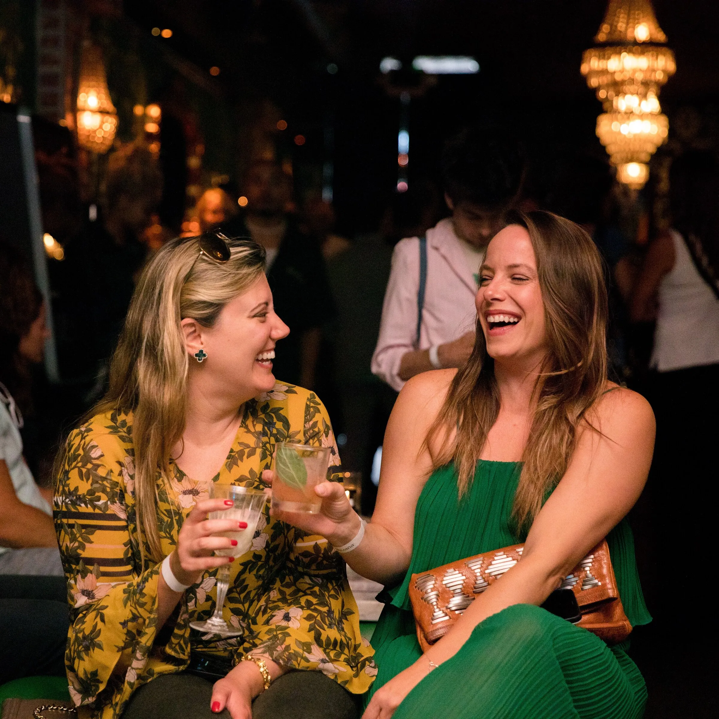 Two guests laughing together with drinks in hand under crystal chandeliers at an evening cannabis cocktail event
