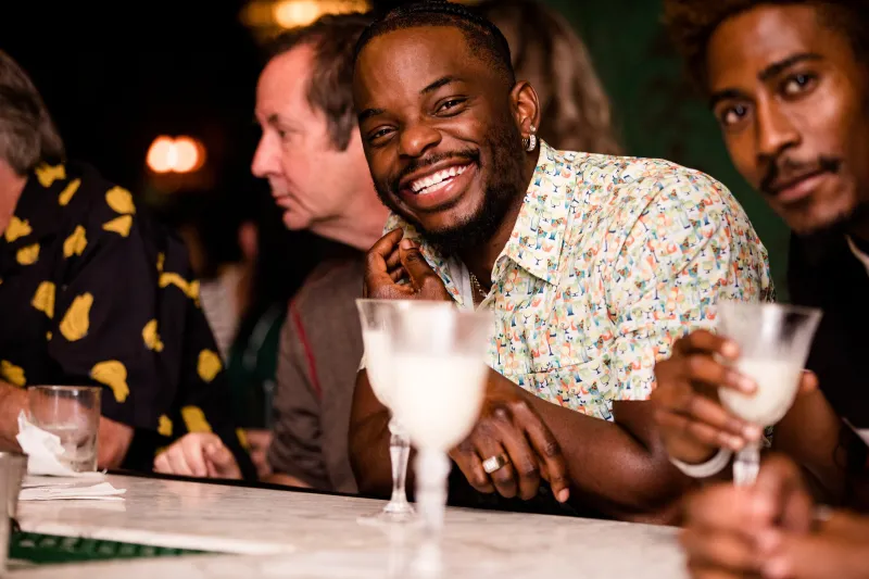 A guest in a patterned shirt grinning at the camera from a marble bar, a cocktail in front of him and friends beside him