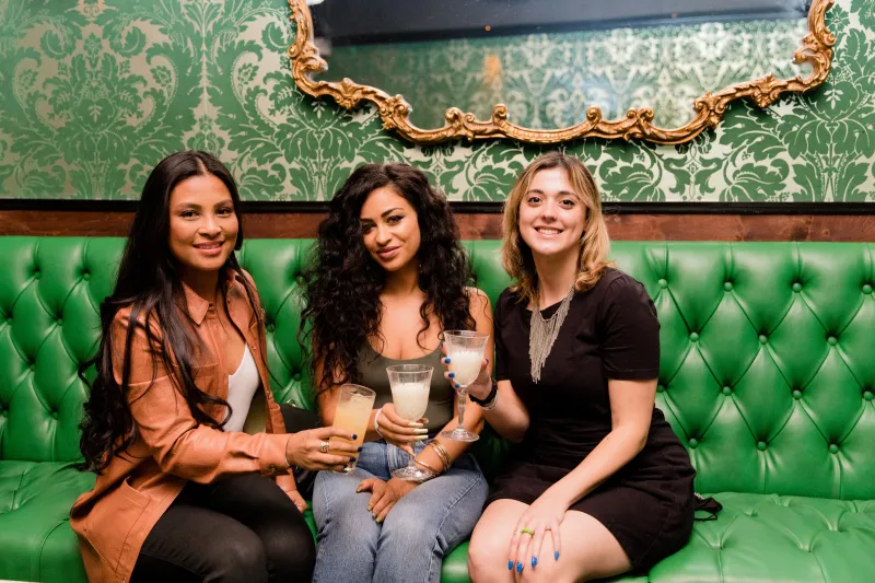 Three friends smiling with cocktails on a tufted green booth under a gilded mirror