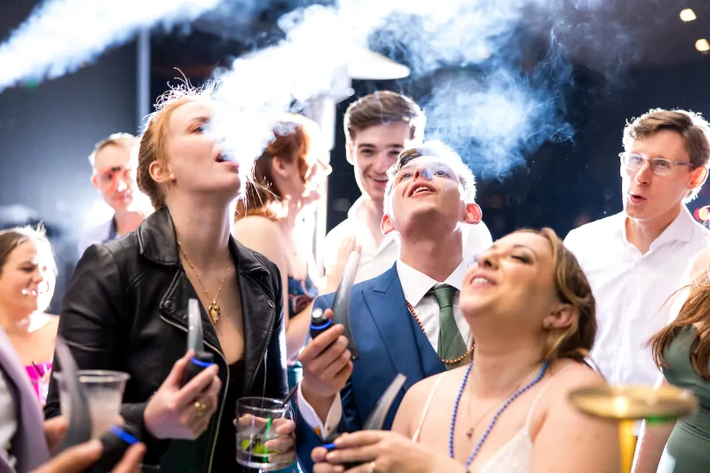 A wedding guest in a leather jacket exhaling a plume of vapor as friends laugh around the cannabis bar