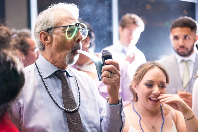 A wedding guest in glow-stick glasses and beads taking a hit from a vaporizer while the bride laughs beside him