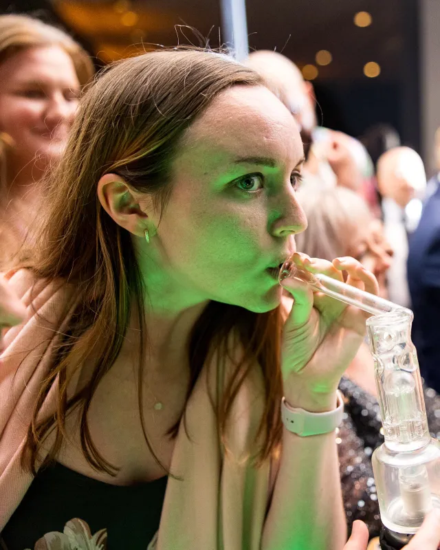 A wedding guest taking a hit from a glass rig at the cannabis bar, lit green, as the party carries on behind her
