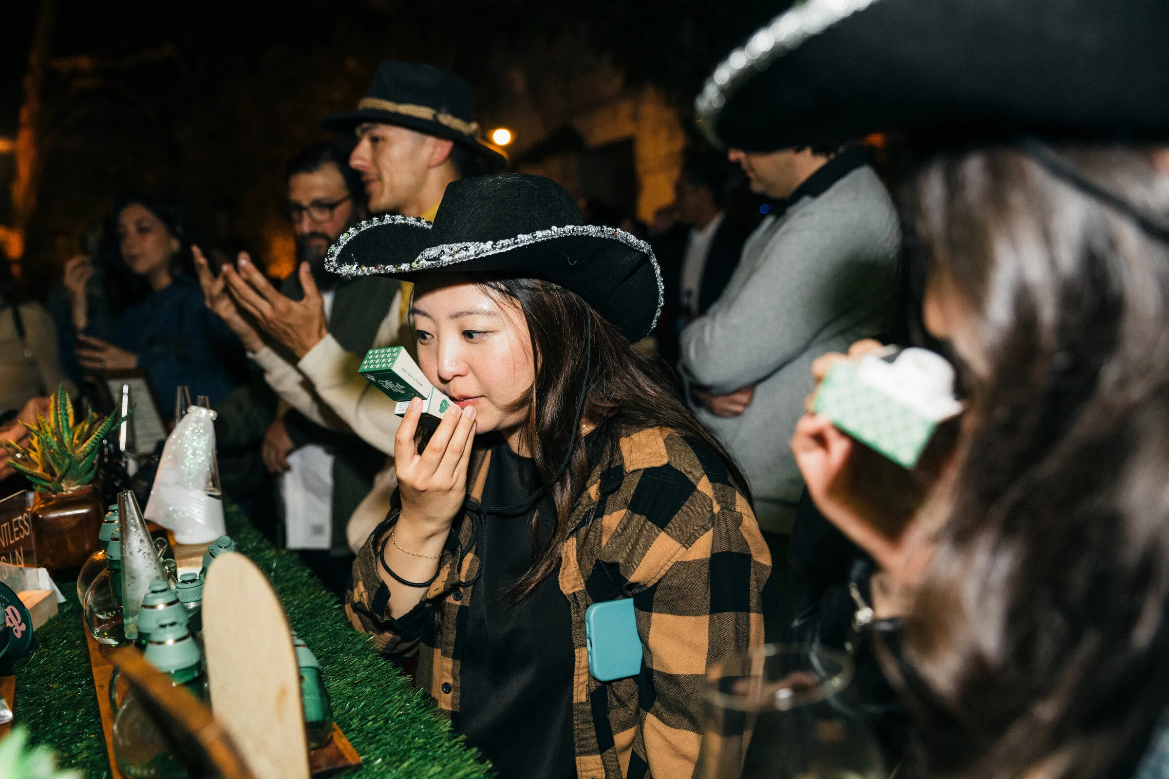 Guest in a plaid shirt smelling a sample at a green cannabis bar, surrounded by guests in black hats at a night party