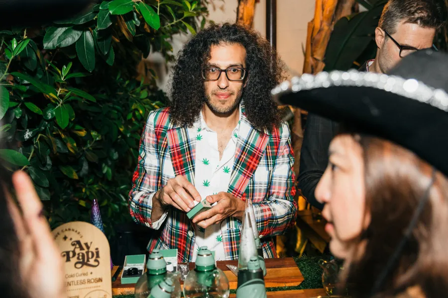 Budtender in a plaid jacket preparing a device behind a cannabis bar with an Ay Papi solventless rosin sign