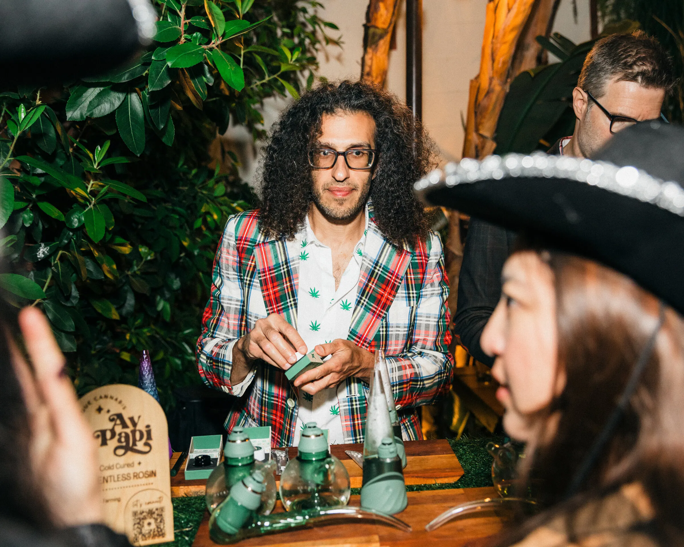 Budtender in a plaid jacket preparing a device behind a cannabis bar with an Ay Papi solventless rosin sign