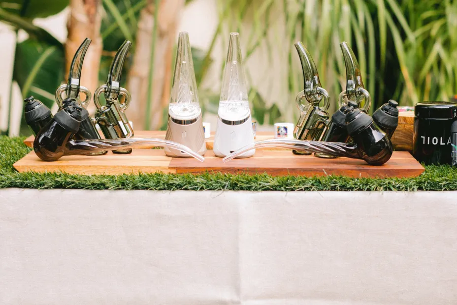 Devices and glassware lined up on a grass-topped cannabis bar in a garden venue, palms behind it