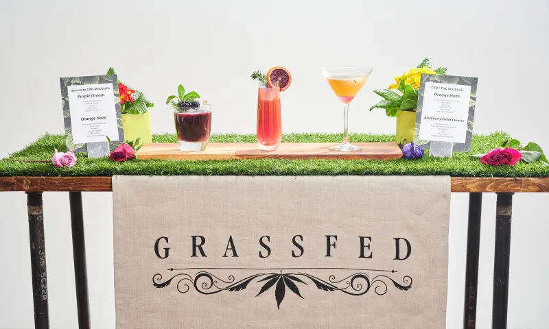 Four CBD and THC mocktails on a grass-topped bar with menu cards and flowers, above a Grassfed table runner