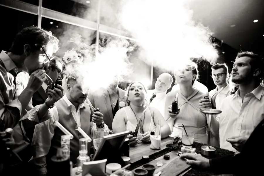 Black and white photo of wedding guests gathered around the cannabis bar, the bride in the center exhaling a cloud of vapor