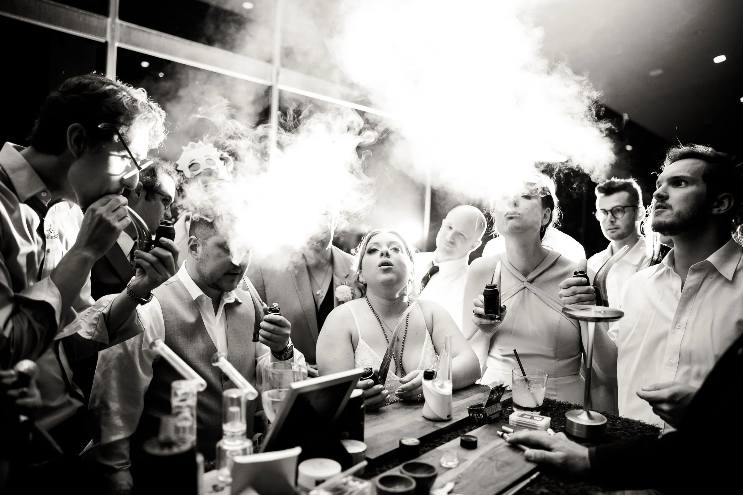 Black and white photo of wedding guests gathered around the cannabis bar, the bride in the center exhaling a cloud of vapor