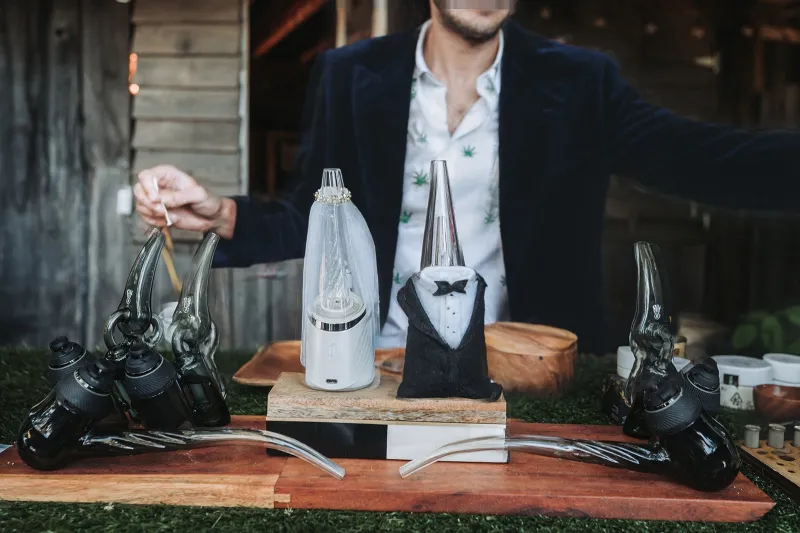 Budtender in a plaid jacket setting up devices on a wood and grass cannabis bar at a rustic outdoor wedding