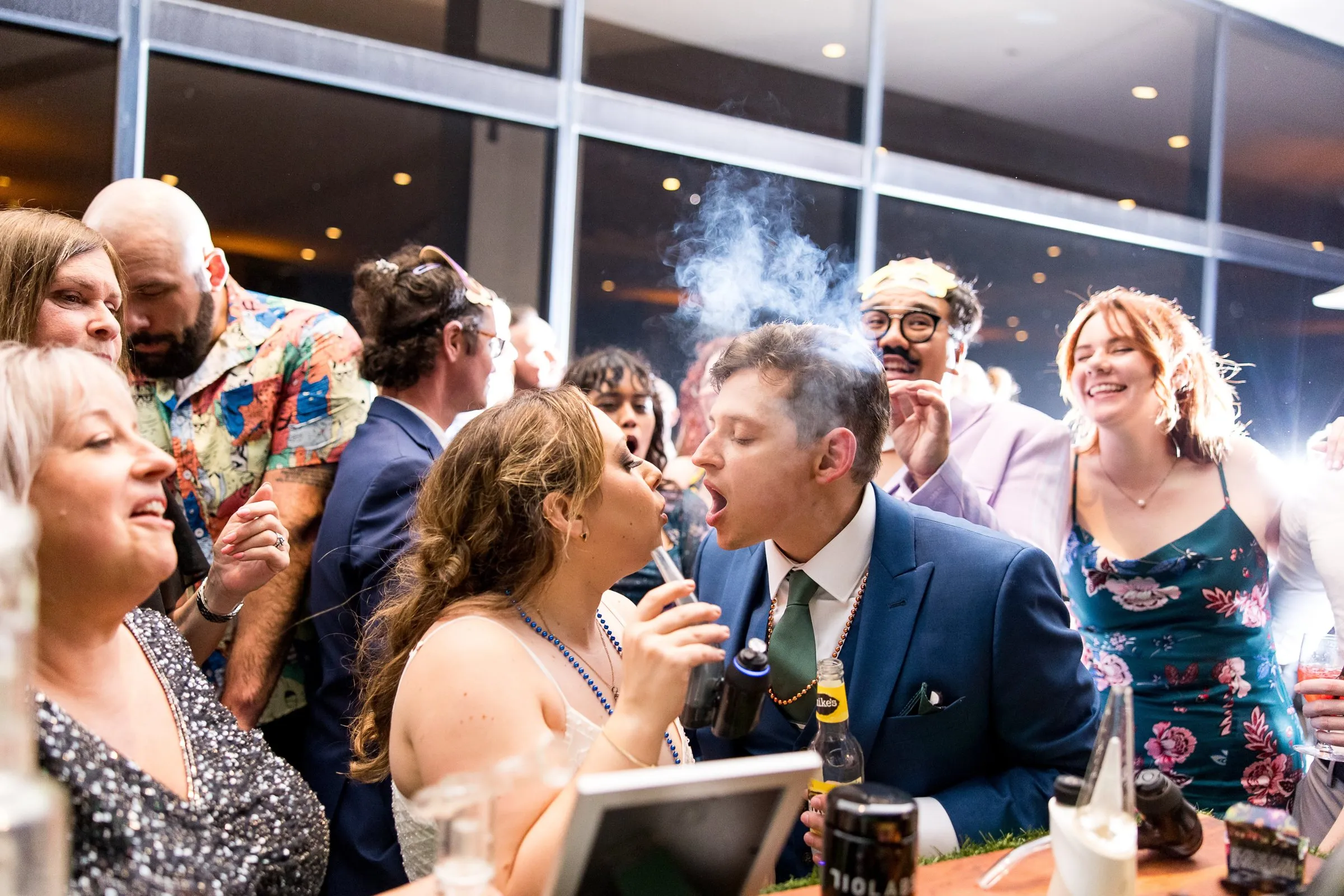 Bride and groom sharing a cloud of vapor at the cannabis bar as laughing guests crowd around them