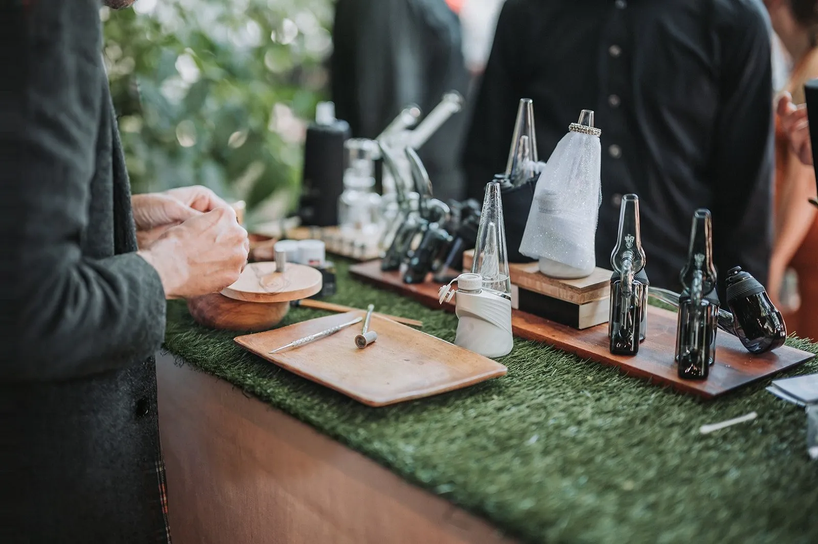 Devices, glassware and wooden trays lined up on a grass-topped cannabis bar at a wedding