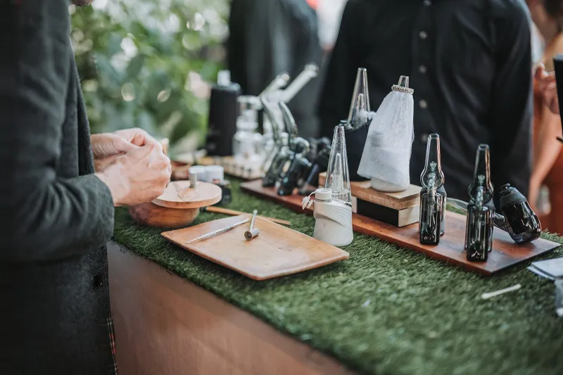 Devices, glassware and wooden trays lined up on a grass-topped cannabis bar at a wedding