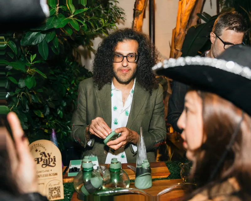 Budtender in a plaid jacket serving guests at a cannabis bar surrounded by tropical plants, an Ay Papi solventless rosin sign on the bar